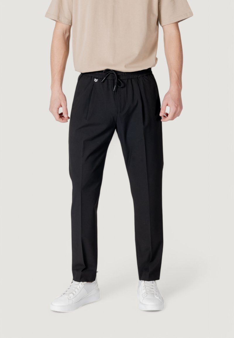 Personne portant un pantalon noir slim-fit avec une ceinture à cordon et des baskets blanches, debout devant un fond clair uni.