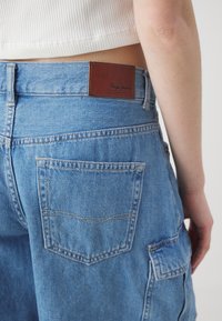 Svetlomodré denimové nohavice s hnedou koženou nášivkou označenou "Pepe Jeans." Obsahujú dvojité zadné vrecká s jemným prešívaním.