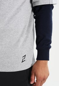 Šedé bavlněné tričko s námořnickými dlouhými rukávy; má černé logo poblíž lemu, které ukazuje na uvolněný design a kontrastní barevné schéma.