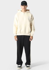 Crème hoodie met een voorzak en een trekkoord, gecombineerd met zwarte sweatpants met zijkanten. Een zwarte pet maakt de outfit compleet.