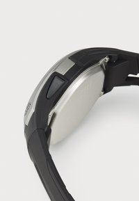 Montre-bracelet avec un strap en caoutchouc noir et un boîtier en métal, dotée d'un dos en argent, d'un bouton latéral et d'accents texturés.
