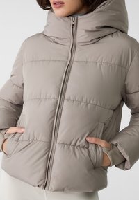 Padded béžová bunda s vysokým golierom, so zipsom vpredu, bočnými vreckami a rebrovanými manžetami. Materiál má hladkú textúru.