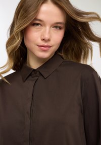 Camicia marrone scuro a maniche lunghe con colletto alla coreana con bottoni, texture liscia e patta frontale. Presenta una leggera lucentezza e una vestibilità sagomata.