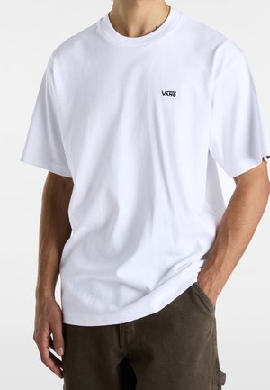 Homme portant un T-shirt Vans blanc uni avec logo sur la poitrine et un pantalon foncé, main dans la poche, sur un fond blanc uni.