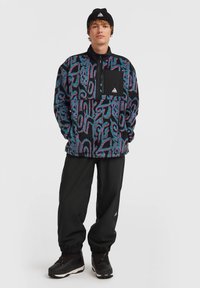 Pull en polaire coloré avec des motifs géométriques en bleu, rose et noir ; équipé d'une poche zippée noire et d'un col haut. Accompagné d'un pantalon noir.