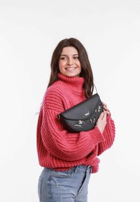 Černá kožená kabelka přes rameno s vý�šivkou motýla, zakřiveným designem a kovovým akcentem. Modelka má na sobě růžový pletený svetr a modré džíny.