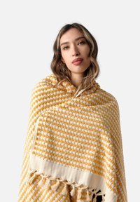 Teksturowany poncho w żółtym i białym kolorze w kratkę, z frędzlami na krawędziach i miękkim, przytulnym wykończeniem.