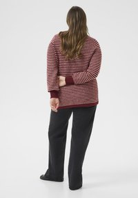 Sweter w kolorze burgundowym, dziergany, z teksturowanym wzorem, z ściągaczami na mankietach i u dołu. Noszony na czarnych spodniach, widziany od tyłu.