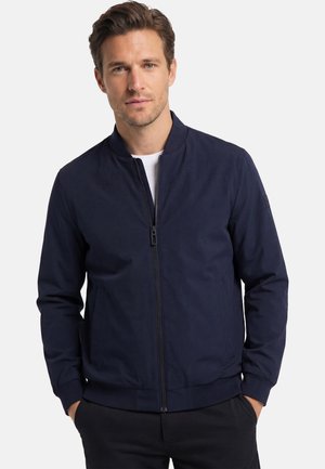 Mann trägt eine marineblaue Bomberjacke mit Reißverschluss über einem weißen Hemd, die Hände in den Taschen, schaut leicht zur Seite vor weißem Hintergrund.