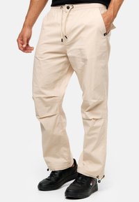 Beige leichte Hose mit elastischem Bund, Kordelzug, Seitentaschen und verstellbaren Bündchen. Kombiniert mit schwarzen Sneakers.