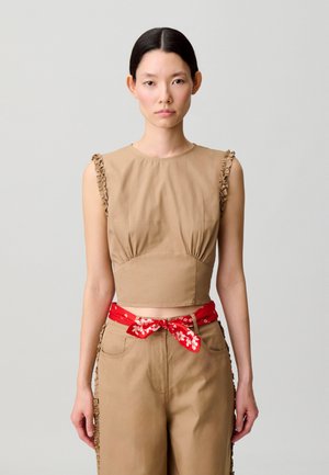 Donna che indossa un crop top beige senza maniche e pantaloni abbinati con volant ai lati, accessoriata con una sciarpa rossa a fiori annodata in vita.