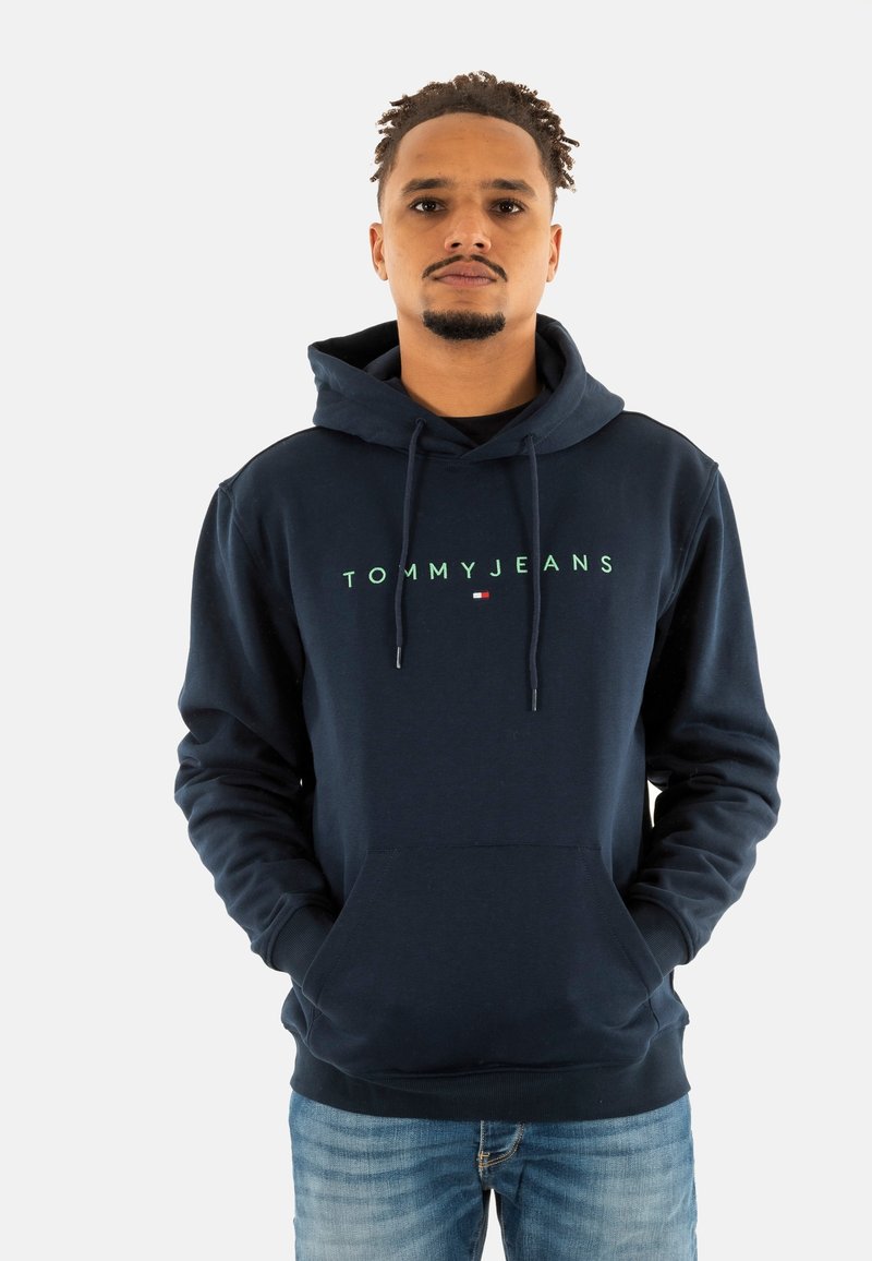 Navyblauwe hoodie van zacht materiaal, met een voorzak en een capuchon met trekkoord. "TOMMY JEANS" logo in lichtgroen over de borst.