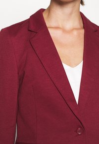 Bordeauxrode blazer met ingesneden revers, enkele knoopsluiting, gestructureerde stof en een getailleerde pasvorm, gecombineerd met een wit V-hals hemd.