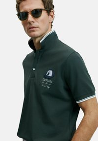 Man met schildpadbril en een donkergroene polo met een wit gestreepte kraag en het "LUFIAN" logo op de borst.