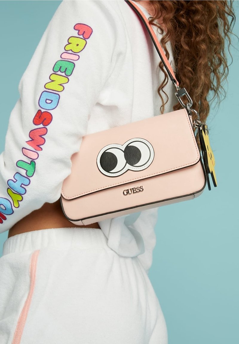 Personne portant une tenue blanche avec le texte coloré FRIENDS WITH YOU sur la manche, portant un sac rose Guess avec un grand design d'yeux de dessin animé.