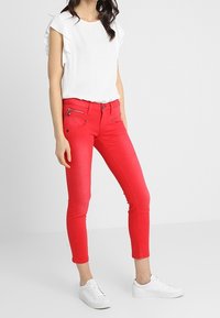 Jeans ajustados rojos con bolsillos frontales con cremallera, combinados con una blusa blanca de manga corta con volantes. Estilizados con zapatillas blancas.
