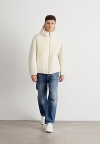 Bijela flis jakna s prednjim zatvaračem i kapuljačom, u kombinaciji s plavim trapericama i bijelim tenisicama, nosi muški model koji stoji.