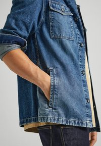 Jeansjacka i mellanblått med knappstängning, bröstficka, uppvikta ärmar och sidfickor. Bärs över en beige topp.