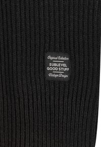 Schwarzer gerippter Strickstoff mit einem rechteckigen Etikett, auf dem in weißen Buchstaben "Original Collection SUBLEVEL GOOD STUFF Vintage Design" steht.