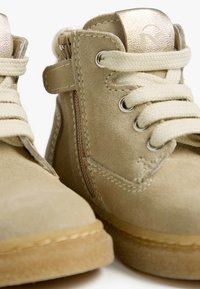 Beige Wildleder-Stiefeletten mit Schnürsenkeln, metallischen Akzenten und Reißverschluss. Hellfarbene Sohle mit Naht am Rand.