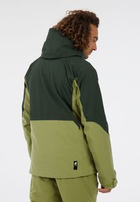 Zelená dvojfarebná bunda s kapucňou, s textúrovaným povrchom a logom na spodnej časti. Horná časť je tmavozelená, dolná časť je svetlozelená.