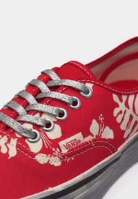 Κόκκινο αθλητικό παπούτσι Vans με λευκό λουλουδάτο σχέδιο, ασημένιες κορδόνια με γκλίτερ και μαύρη σόλα που δείχνει ελαφριά φθορά και ετικέτα της μάρκας στο πλάι.