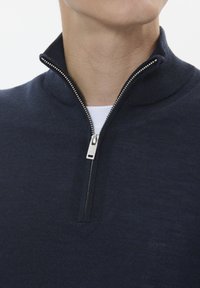 Mornarska zip-jaka z visokim ovratnikom, ki ima srebrno zadrgo in rebrasto teksturo. Material deluje mehek in pleten.