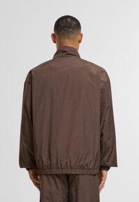 Moški, oblečen v ohlapen rjav plašč in ujemajoče se hlače, stoji obrnjen stran od preproste svetle ozadja.