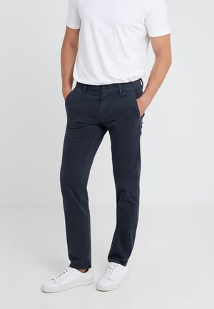 Mężczyzna ubrany w granatowe spodnie slim fit, białą koszulę z krótkim rękawem oraz białe tenisówki, stojący na jasnoszarym tle, z rękami w kieszeniach.