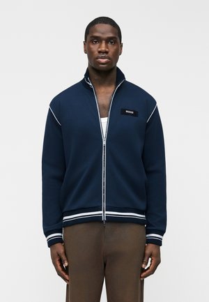 Moški, oblečen v mornarsko modro jakno z zadrgo in belimi poudarki ter rjave hlače, stoji pred enobarvnim svetlim ozadjem.