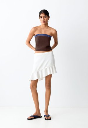 Žena u smeđem topu bez naramenica s plavim obrubom, asimetričnoj bijeloj suknji s faltenom na rubu i crnim japankama, stoji na bijeloj pozadini.