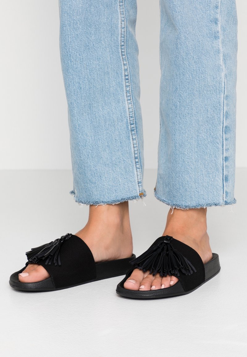 Pieds portant un jean bleu clair à ourlet effiloché et des sandales noires à bout ouvert avec des détails à pompons sur le dessus.