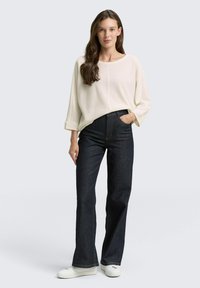 Witte, gestructureerde gebreide top met brede mouwen; donkerblauwe flairjeans; witte sneakers; model poseert met één hand in de zak.