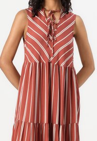 Robe sans manches rouge rouille avec des rayures horizontales et diagonales crème. Présente un col noué et une jupe évasée plissée. Texture de tissu lisse.