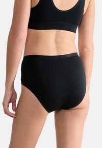 Vrouw in zwarte naadloze high-waist onderbroek en bijpassende zwarte sportbra, zichtbaar van achteren tegen een effen achtergrond.