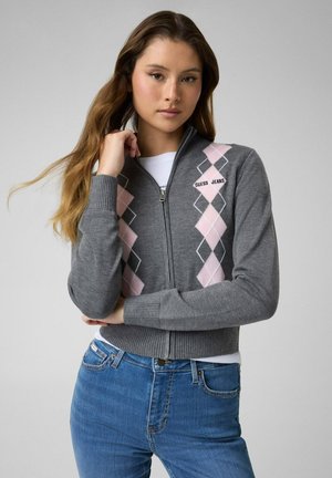 Junge Frau trägt einen grauen Zip-up-Pullover mit pinkem Argyle-Muster und blauer Jeans, steht vor einem einfarbigen hellen Hintergrund.