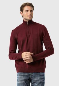 Bordeauxroter Reißverschluss-Pullover mit hohem Kragen und gerippten Bündchen. Strickstruktur, zeigt ein dezentes Muster, getragen über hellblauen Jeans.