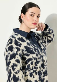 Leopardmönstrad cardigan i krämfärgad och marinblå, texturerad stickad fabric, svarta knappar, lager över en marinblå skjorta med krage.