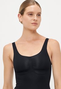 Young woman with light skin and blonde hair in a sleek bun wearing a black sleeveless fitted top, looking slightly right on a plain background.