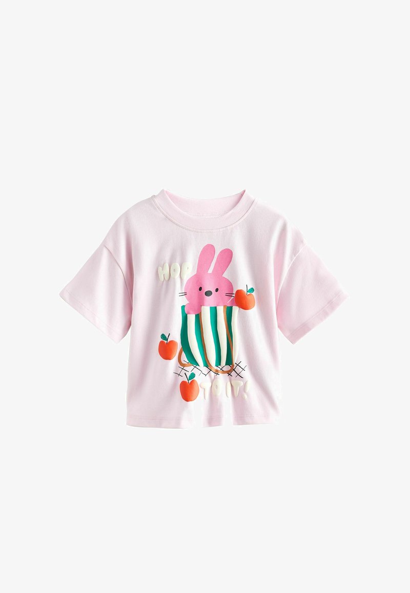 Lichtroze shirt met korte mouwen met een ontwerp van een roze konijntje in een groen gestreepte mand, omringd door drie rode appels.