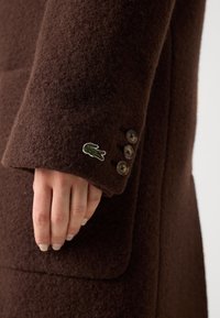 Cappotto in lana marrone con finitura testurizzata, tre bottoni scuri sui polsini e un piccolo logo di coccodrillo ricamato.