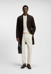 Mörkbrun kappa, krämfärgad tröja, ljuskrämfärgad byxor och bruna loafers. Svart väska med rem hålls i handen. Neutral bakgrund.