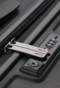 Valigia nera con cerniere metalliche, una serratura a combinazione e il nome del marchio "BRICS" impresso sui tiranti delle cerniere argentate.