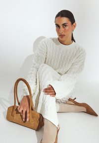 Donna con capelli scuri in un vestito a maglia bianca, collant trasparenti beige e décolleté in suede marrone, mentre tiene una borsa in suede marrone.