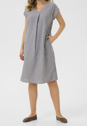 Femme portant une robe grise, courte aux genoux, à manches courtes, avec des rayures verticales blanches et des chaussures plates marron, main dans la poche, debout sur fond blanc.