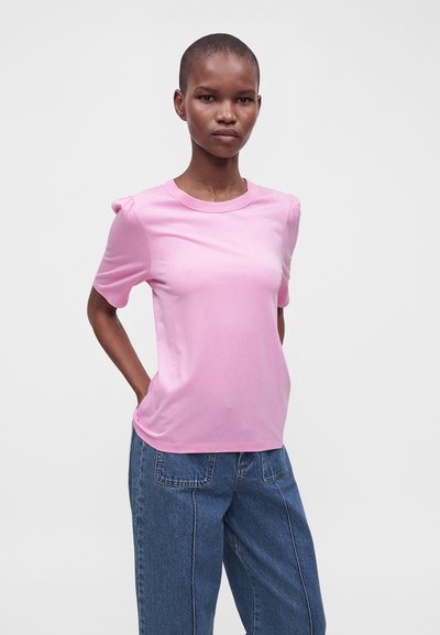 Jeune personne aux cheveux courts portant un t-shirt rose clair à manches courtes et un jean bleu large, debout les mains dans le dos.