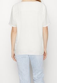 Bílý krátký rukáv top s volným střihem, který má kulatý výstřih a texturovanou látku. Zadní pohled ukazuje jednoduchý, nezdobený design.