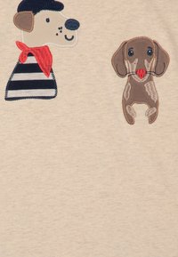 Besticktes T-Shirt aus beigem Stoff mit einem Hund in einem gestreiften Hemd und einem roten Schal sowie einem braunen, gefleckten Hund mit einem roten Ball.