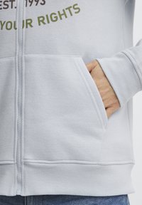 Svijetlo siva zip-up majica s duplim prednjim džepovima, rebrastim rukavima i mekim teksturom. Prednja strana ima višebojni natpis.