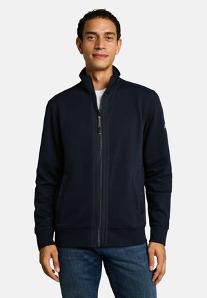 Mann mit einer dunkelblauen Jacke mit Reißverschluss über einem weißen Hemd und blauen Jeans, steht vor einem einfarbigen hellen Hintergrund.
